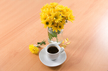 xícara de café com flor amarela ao lado e vaso de flor no fundo de madeira.