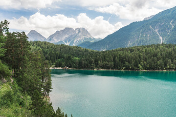 Naklejka na ściany i meble An emerald green Blindsee lake surrounded by forest and mountains, with hiking path on a side