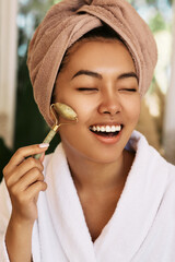 Happy Asian woman with closed eyes using green jade roller for face massage, close-up