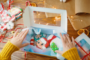 Women's hands open gift box with Christmas cookies. Gingerbread with icing. Festive sweets for children in form of pig, mittens, pine tree and snowflakes. New Year's composition on wooden table.