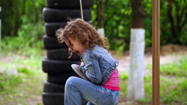 Little curly girl 4 years old in a denim suit rides a swing in summer