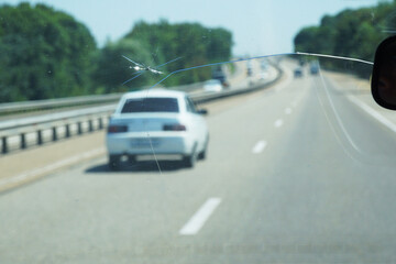 A crack in the windshield from a stone that flew in from under the wheels of a car against the background of a road and another car.
