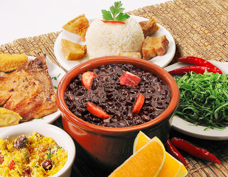 Feijoada completa, comida típica brasileira com arroz, feijão preto, linguiça, bacon, torresmo, farofa, couve e laranja, em fundo de esteira de palha.