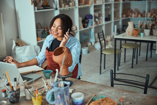 Cheerful female potter talking on cellphone in pottery workshop