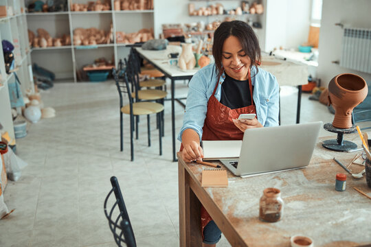 Joyful woman using laptop and smartphone in pottery workshop - Powered by Adobe