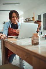 Cheerful woman using smartphone and laptop in pottery workshop