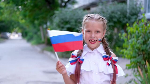 A schoolgirl stands in the street and waving the Russian flag. Happy little girl with face painting of Russian symbols. Day of the Russian flag.