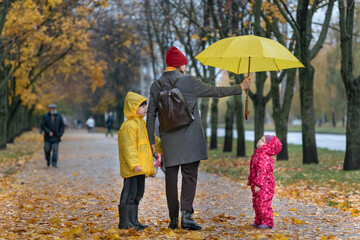 Mother with two children is walking in the autumn park. Mom holds a yellow umbrella over her daughter.