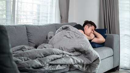 Asian man is lying with blanket on couch at home to rest when having high fever, headache and using