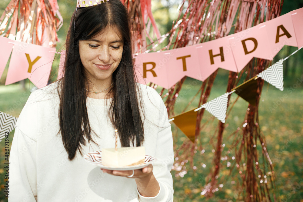 Wall mural Stylish happy woman in party hat holding piece of birthday cake with burning candle and making a wish on background of pink happy birthday garland. Celebrating birthday at picnic party outdoor