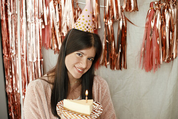 Stylish happy woman in party hat holding piece of birthday cake with burning candle on background...