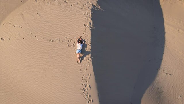 Woman lie on wavy sand dunes in desert landscape at golden sunset light and wind.