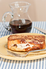 front view, close up, of, freshly baked, maple, bacon, stuffed, french toast, on a round, yellow plate with artisan knife and fork with a glass pitch filled with syrup on a blue and white table cloth