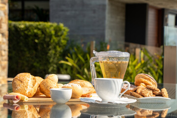 afternoon tea served on a transparent table, with bread, jam, cheese and ham and donuts with a clear sky and green plants in the background
