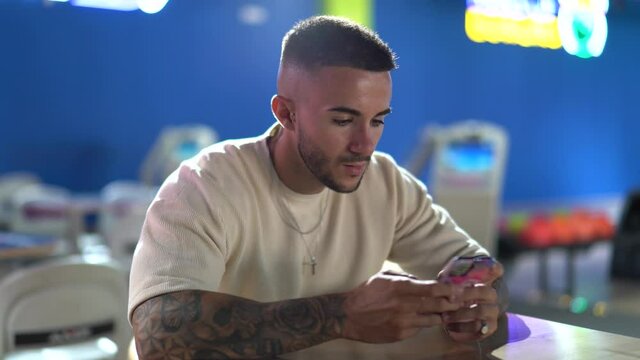 A young Spanish handsome man with tattoos texting on his phone in a bowling arena shot in 4K