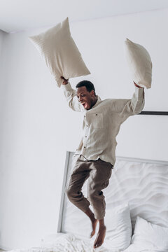 Joyful young African-American man in casual clothes has fun jumping and throwing pillows on large bed at home in sunny morning