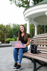 A smiling European dark-haired girl in modern clothes, sitting on a park bench in the summer and looking away with a laptop, a bag, notebooks, and a pen.