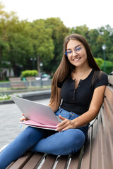 Obraz premium Freelance, business, technology, and Internet concept — A smiling European dark-haired women sitting on a park bench in the summer with a laptop, pen, and notebooks looking into the camera.