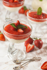Several glasses with traditional creamy pudding dessert panna cotta with peppermint and strawberry jam surrounded fresh strawberries on a marble surface