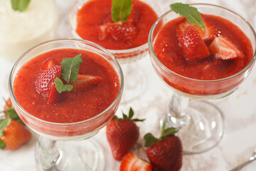 Several glasses with traditional creamy pudding dessert panna cotta with peppermint and strawberry jam surrounded fresh strawberries on a marble surface