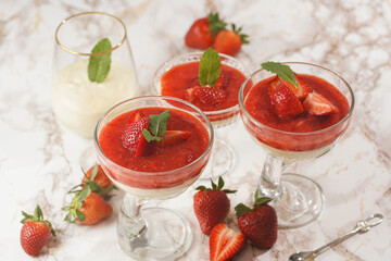 Several glasses with traditional creamy pudding dessert panna cotta with peppermint and strawberry jam surrounded fresh strawberries on a marble surface