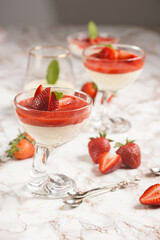 Several glasses with traditional creamy pudding dessert panna cotta with peppermint and strawberry jam surrounded fresh strawberries on a marble surface