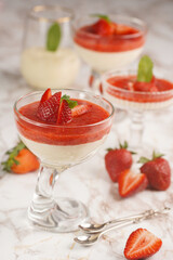 Several glasses with traditional creamy pudding dessert panna cotta with peppermint and strawberry jam surrounded fresh strawberries on a marble surface