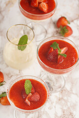Several glasses with traditional creamy pudding dessert panna cotta with peppermint and strawberry jam surrounded fresh strawberries on a marble surface