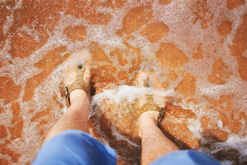 Men's feet in sandals on beach. Man in jeans shorts stands and looks down at his feet in sand that...