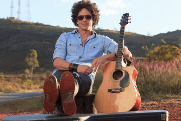 man musician guitarist with curly hair and his guitar and case sitting by the roadside on red ground in mountain summer.