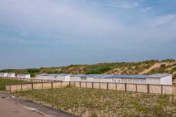 Knokke-Heist, Flanders, Belgium - August 5, 2021: Row of white cabins set in dunes and shielded from Nord Sea under blue sky. Boardwalk in front.