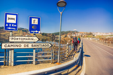 Pilgrims Hiker Backpackers Crossing Bridge over Lake Water to Portomarin, Spain, along the Way of...