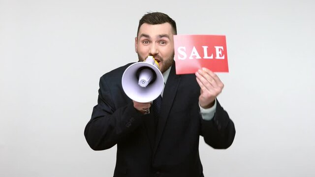 Excited bearded businessman in black official style suit screming at megaphone and showing card with sale inscription, announcing discounts. Indoor studio shot isolated on gray background.