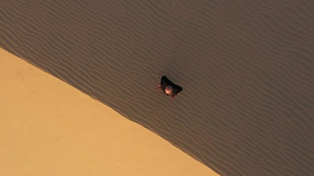 Old man in black is engaged in kung fu in the dunes at sunset. a lot of sand, golden sunset light. sports training. handsome man meditating. drone goes up. aerial top view to sound dunes on sunset