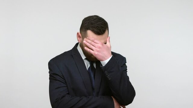 Bearded businessman in black official style suit doing facepalm gesture, touching head with sorrow regret expression, desperate about trouble. Indoor studio shot isolated on gray background.