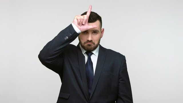 Upset sad bearded businessman in black official style suit standing showing loser gesture, upset about dismissal, making L sign on forehead. Indoor studio shot isolated on gray background.