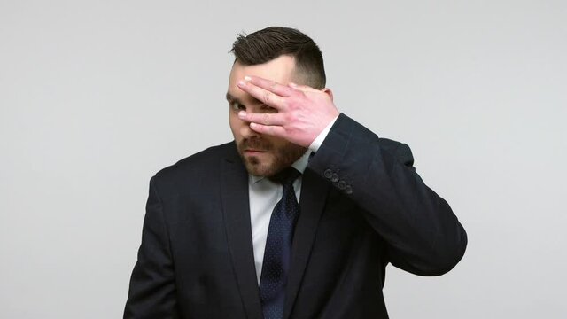 Bearded businessman in black official style suit covering eyes with palm, peeking through fingers, looking with suspicion, shy to watch forbidden event. Indoor studio shot isolated on gray background.