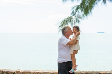 Happy Asian family on summer travel vacation. Grandfather carrying and playing with grandchild girl on the beach. Senior man with granddaughter enjoy and having fun outdoor activity lifestyle together
