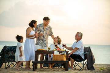 Happy Asian family on holiday vacation. Group of multi generation family little girl with parents and grandparents relax and enjoy with dinner party and dancing together on the beach at summer sunset.