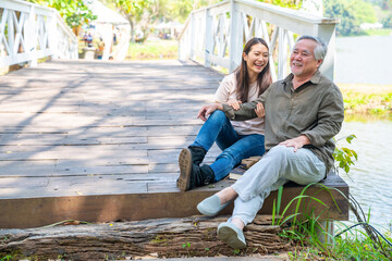 Asian woman and senior man father sitting and talking together on wooden bridge in park. Elderly retired male relax and enjoy outdoor activity lifestyle with his daughter. Family relationship concept