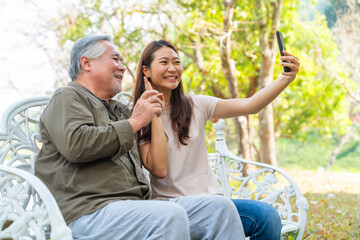 Happy Asian family senior man sitting on bench in the park and using smartphone taking selfie together with granddaughter. Elderly retirement male relax and enjoy outdoor activity with daughter.