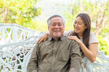 Asian woman caring and hugging senior man grandfather sitting on outdoor chair in the park. Elderly retired male relax and enjoy outdoor activity together with daughter. Family relationship concept