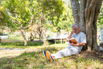 Naklejka na ściany i meble Asian senior man sitting on grass under the tree reading a book in park. Retirement elderly male relax and enjoy outdoor leisure activity in summer day. Older health care and illness recovery concept.