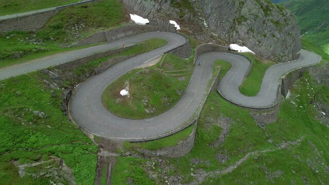 AERIAL: Bicycle rider crosses the Gotthard mountain pass in Switzerland on a cloudy summer day. Flying above an active male tourist as he rides a bicycle up a switchback road in Swiss mountains.