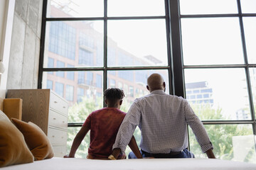Rear view of Black father and son sitting on bed in front of window