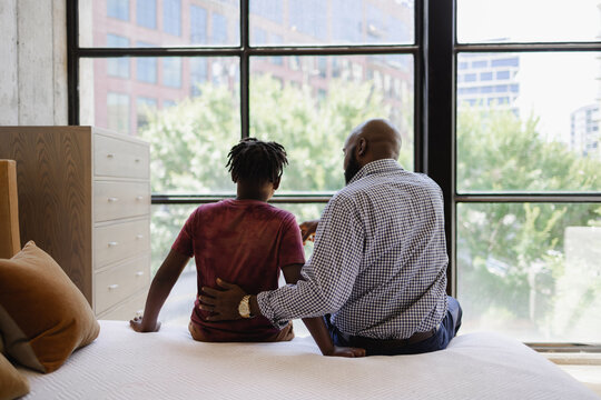 Rear view of father and son sitting on bed in front of window