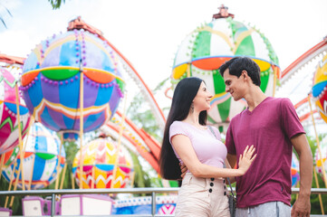 Handsome young man smiling and holding woman by waist standing in theme park while looking at girlfriend with love