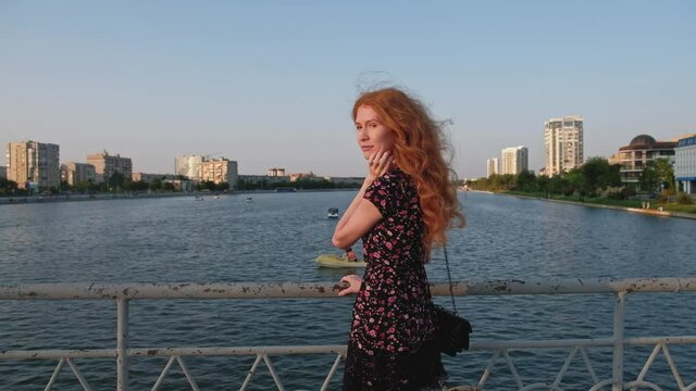 A redheaded lady wearing summer dress looking at view rear view from bridge over river in Astrakhan Russia.