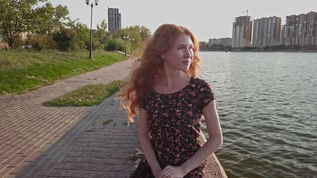 Red haired lady in short summer dress is sitting on the weathered concrete parapet of river side with construction side on background, she looks relaxed and full of promises. Copyspace.