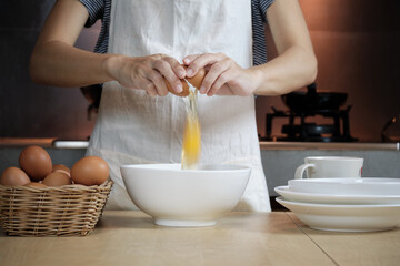 Close-up front view footage, a female cook in a white apron is cracking an egg into a cup to prepare a meal on a wooden table in the home's kitchen. Eating egg yolks is a healthy breakfast.